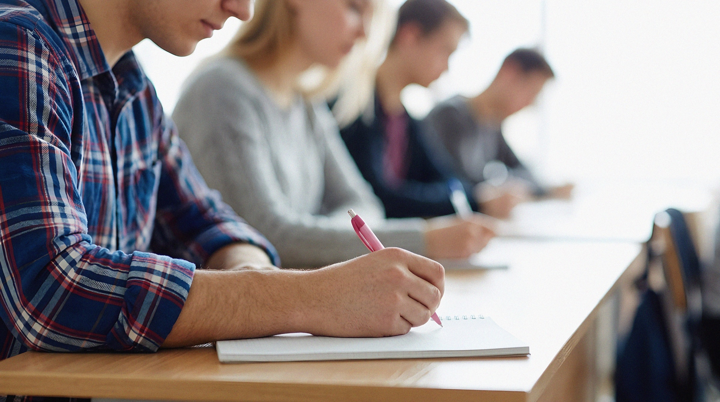 Person in a plaid shirt writing in a notebook while others write nearby.